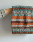 A back shot of Wandrian, a colourwork cowl knit using hand dyed yarn. It is looped over the hand of a white person wearing a grey knitted cardigan and hanging so you can clearly see the colourwork design. The cowl design features abstract patterns of flowers and branches alongside some stripes. The colours used are very autumnal, a lovely teal complimented with a soft grey, a burgundy and a rust orange.