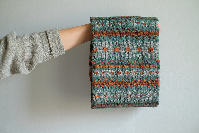 A front shot of Wandrian, a colourwork cowl knit using hand dyed yarn. It is looped over the hand of a white person wearing a grey knitted cardigan and hanging so you can clearly see the colourwork design. The cowl design features abstract patterns of flowers and branches alongside some stripes. The colours used are very autumnal, a lovely teal complimented with a soft grey, a burgundy and a rust orange.