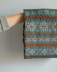 A front shot of Wandrian, a colourwork cowl knit using hand dyed yarn. It is looped over the hand of a white person wearing a grey knitted cardigan and hanging so you can clearly see the colourwork design. The cowl design features abstract patterns of flowers and branches alongside some stripes. The colours used are very autumnal, a lovely teal complimented with a soft grey, a burgundy and a rust orange.
