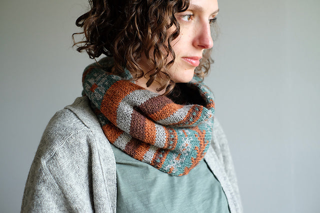 Wandrian, a colourwork cowl knit using hand dyed yarn is being modelled by a white woman with mousy coloured curly hair wearing a grey knitted cardigan. The cowl design features abstract patterns of flowers and branches alongside some stripes. The colours used are very autumnal, a lovely teal complimented with a soft grey, a burgundy and a rust orange