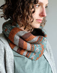 Wandrian, a colourwork cowl knit using hand dyed yarn is being modelled by a white woman with mousy coloured curly hair wearing a grey knitted cardigan. The cowl design features abstract patterns of flowers and branches alongside some stripes. The colours used are very autumnal, a lovely teal complimented with a soft grey, a burgundy and a rust orange