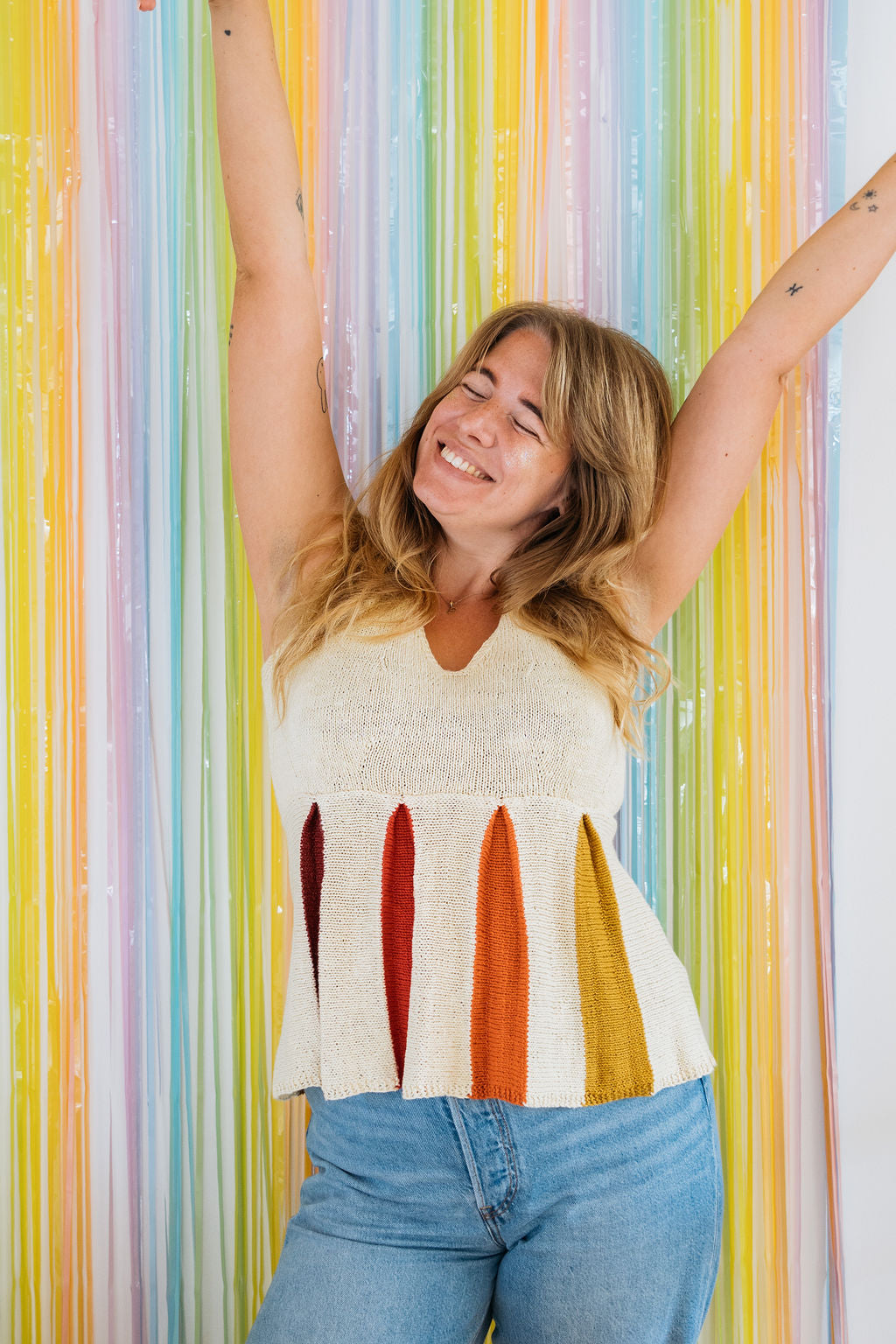 A woman in a colorful knit top and high-waisted jeans poses joyfully against a backdrop of colourful streamers. The knitted top is white with a peplum bottom which has colourful folds.