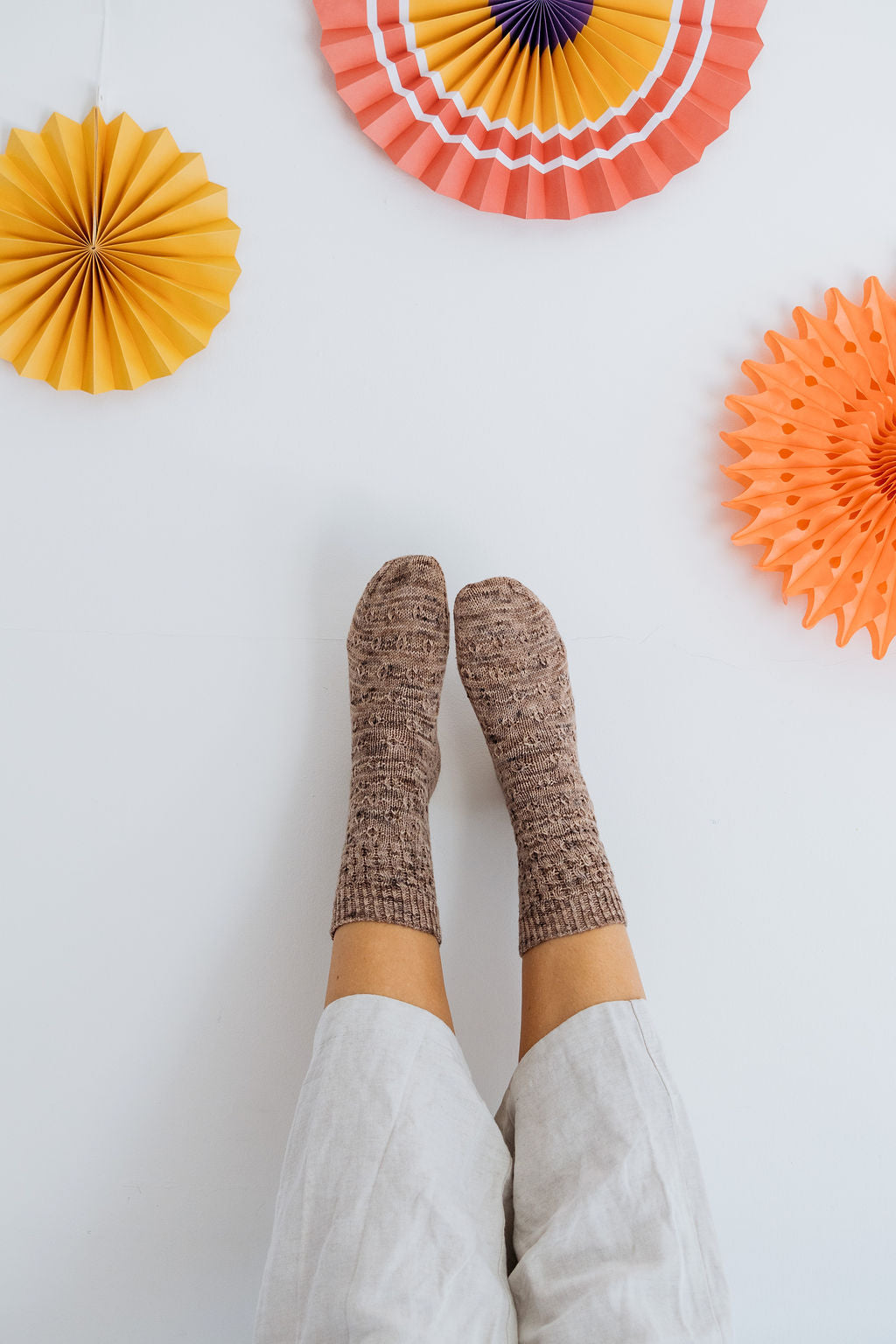 A close up shot of a persons legs held up against a wall. They are wearing white trousers and a speckled pair of handknit socks. The wall is adorned with colourful paper decorations.