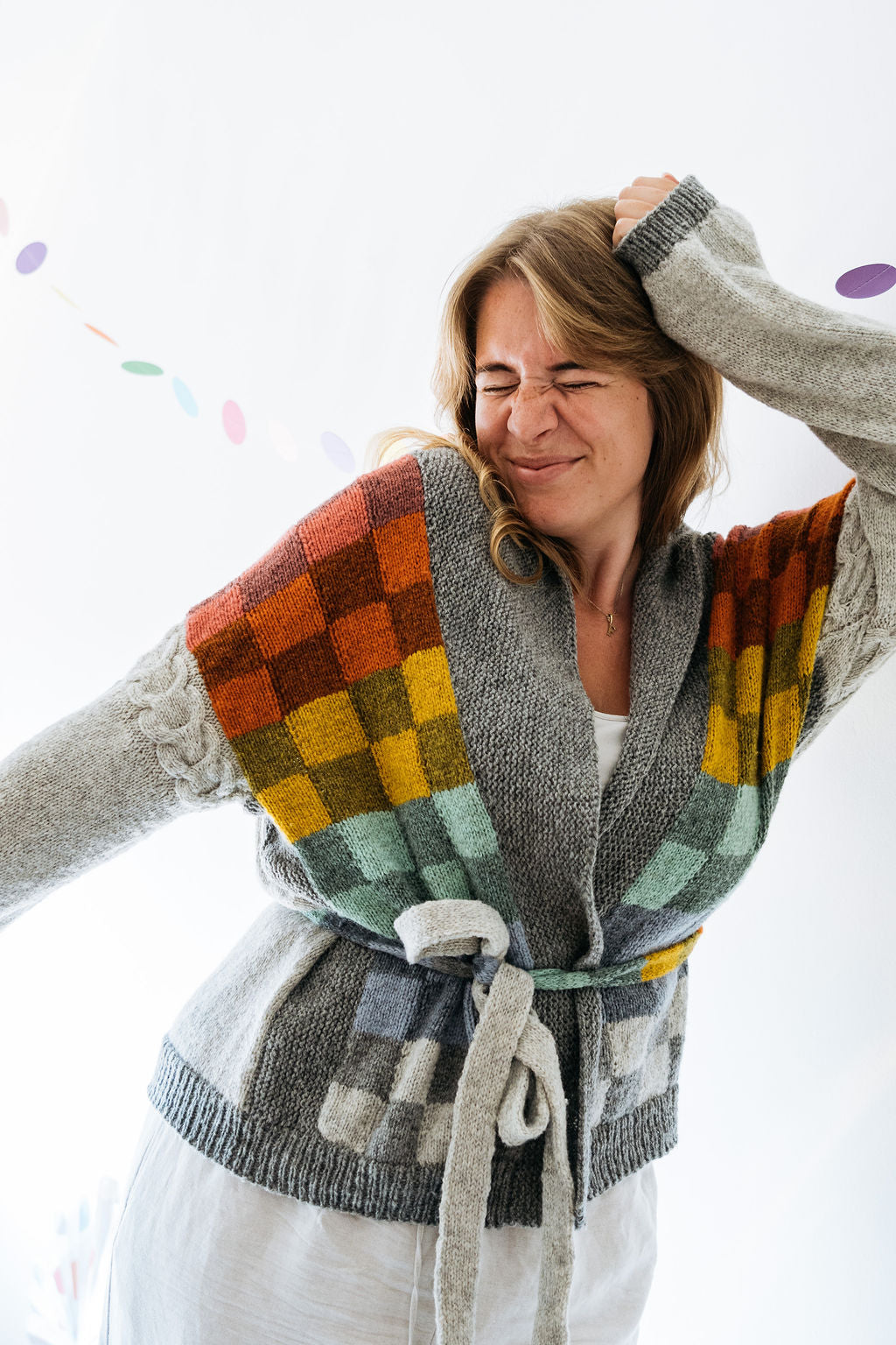 A woman in a white dress wears a grey handknitted cardigan that features colourful checkered colourwork in two panels on the front.