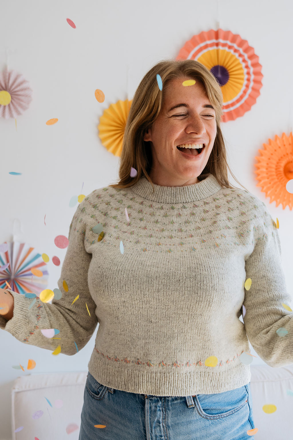 A woman in a handknitted sweater with cute confetti like detailing across the yoke stands amidst colorful confetti, with festive paper decorations in the background, celebrating joyfully.