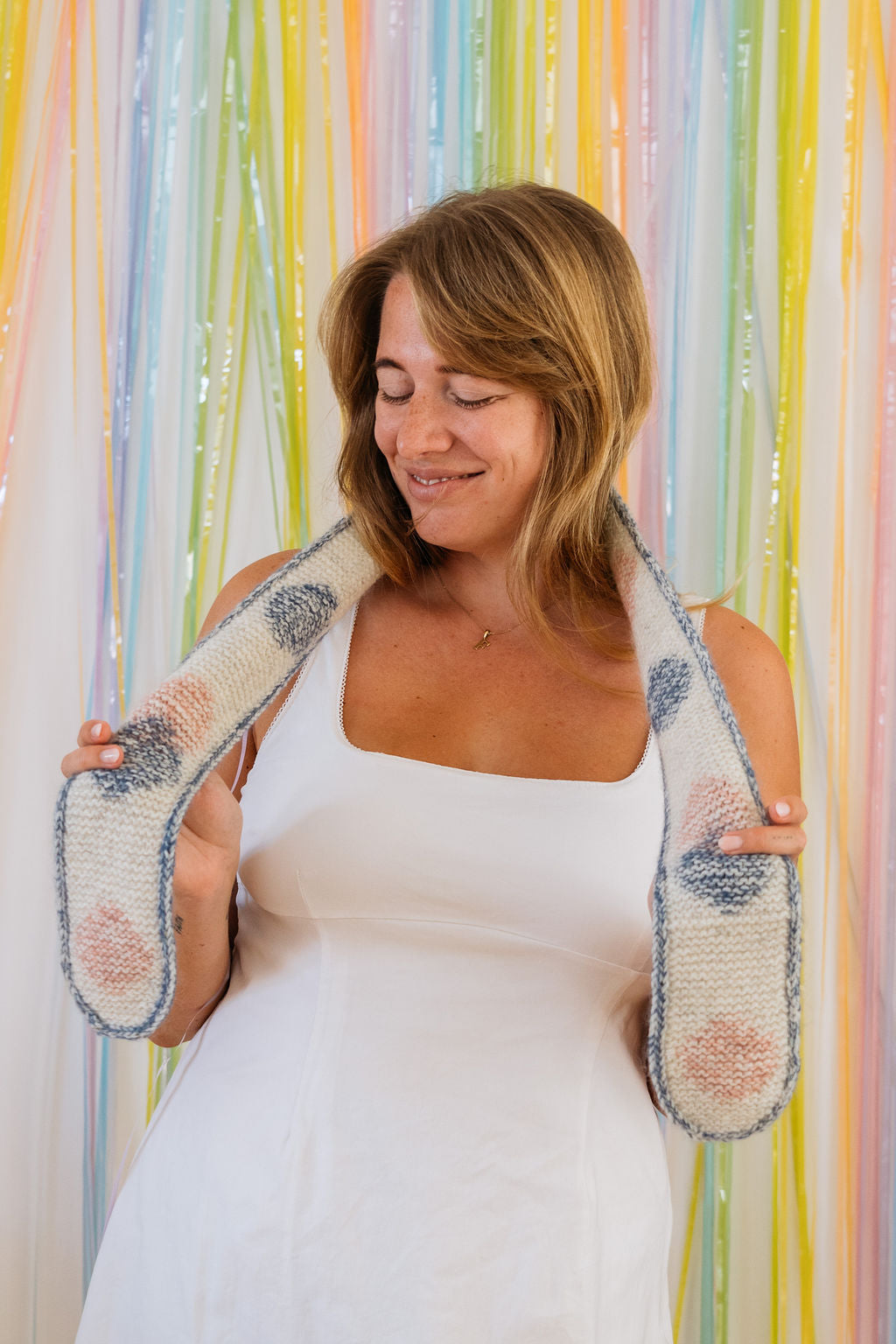 A woman in a white dress wraps a short, hand knitted scarf around the back of her neck. She is holding each end and looking down smiling. Behind her is a backdrop of pastel streamers.