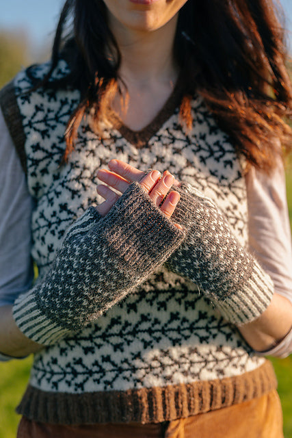 a torso shot of marina. She is wearing a colourwork handknit vest with branch like motifs, but the focus is on her hands held in front of her chest in a pair of colourwork mitts knit in sheepy shades. 