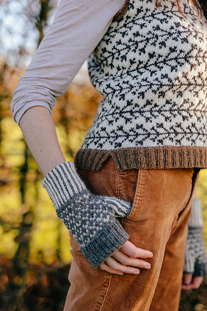 a torso shot of marina. She is standing slightly to the side with her thumb hooked in her pockey wearing a handknit tank top with branch like colourwork and a pair of colourwork mitts knit in sheepy shades. 