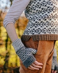 a torso shot of marina. She is standing slightly to the side with her thumb hooked in her pockey wearing a handknit tank top with branch like colourwork and a pair of colourwork mitts knit in sheepy shades. 