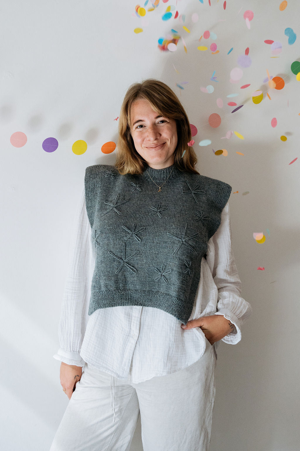 A person wears a gray hand knitted vest with floral embroidered detailing over a white shirt. She is smiling directly at the camera and there is colourful confetti and circular decorations in the background.