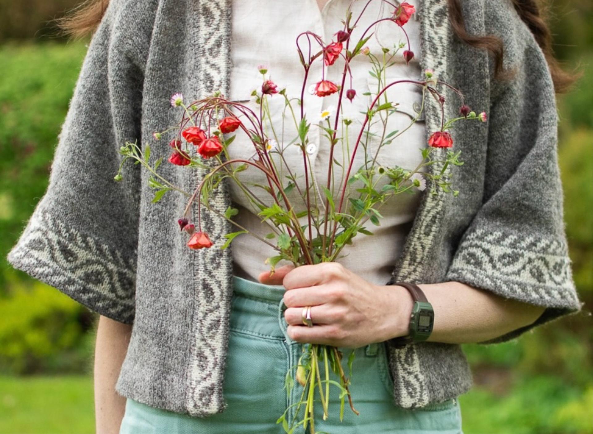 A close up image of marina, a white woman holding a bunch of wild flowers. The image is centred on her hand holiding the flowers and you can see the details of the knitting cardigan she is wearing with colourwork along the sleeve edges and front edges