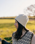 Marina, a white woman with brown hair is standing outside and pictured looking to the side. She is wearing a handknit colourwork vest, but the focus is on the white felted knit hat she is wearing. 