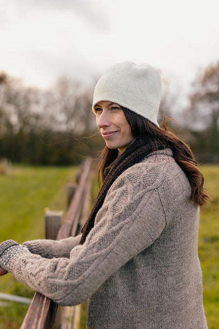 Marina, a white woman with brown hair is standing outside with her arms resting on a fence. She is wearing a cosy grey handknit cardigan, a dark brown scarf and a felted knit hat. She is looking off in the distance with a smile on her face. 