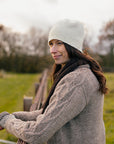 Marina, a white woman with brown hair is standing outside with her arms resting on a fence. She is wearing a cosy grey handknit cardigan, a dark brown scarf and a felted knit hat. She is looking off in the distance with a smile on her face. 
