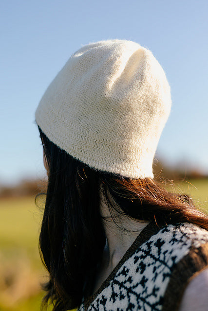 The back view of Marina, a white woman, wearing a creamy white hand-knit hat with a subtle herringbone texture around the brim. Blue sky and countryside fields are blurred in the background.