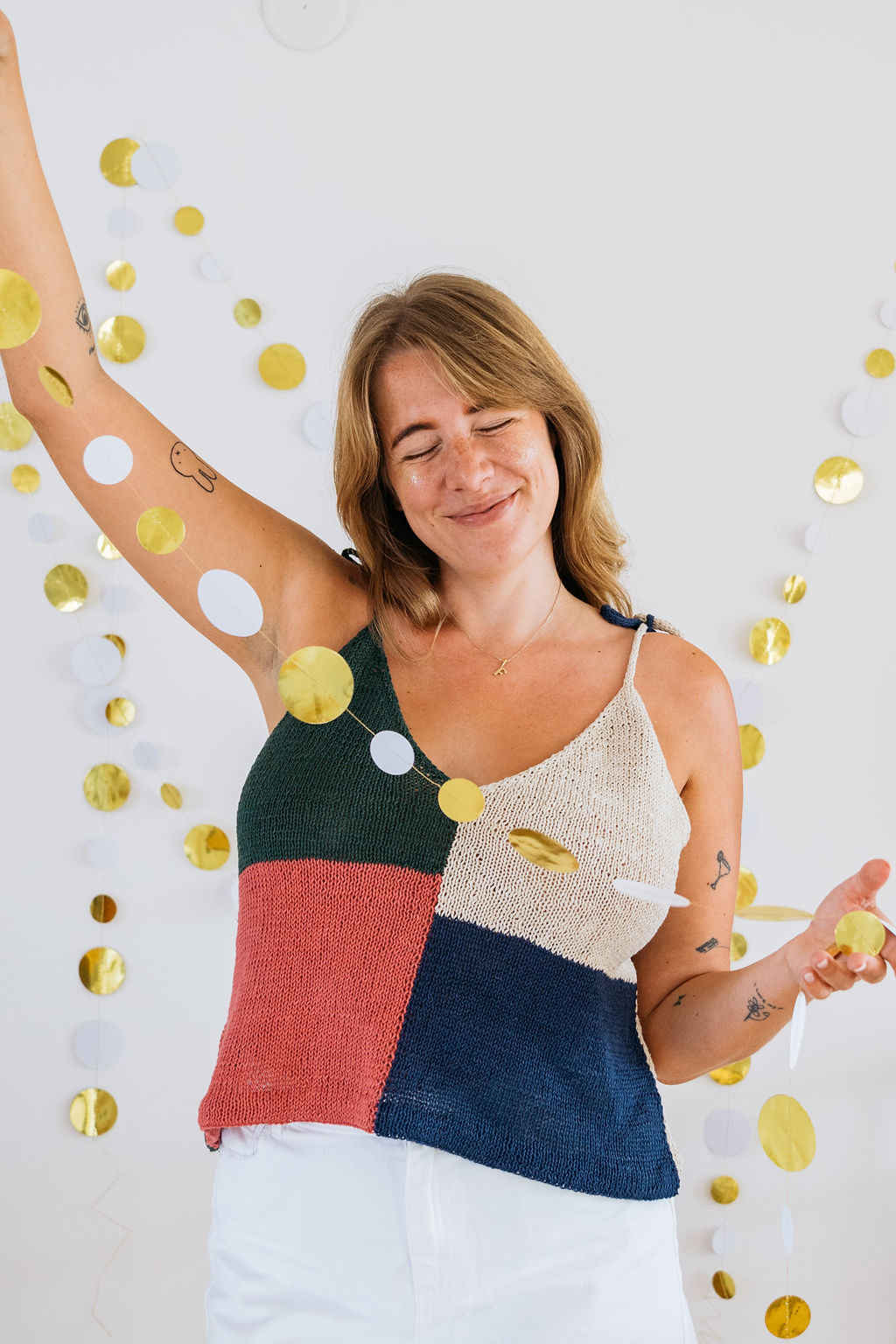 A woman in a colorful, patchwork knit top raises her arm, surrounded by gold and white decorative circles hanging on a white wall.