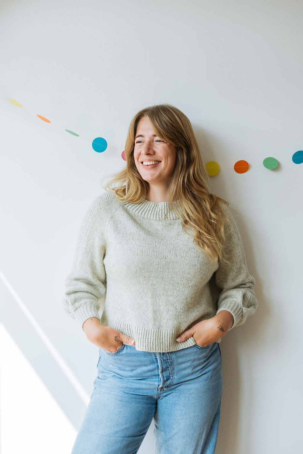 A woman with long, wavy hair wears a cosy light hand knitted sweater and high-waisted jeans. She is posing against a wall decorated with colorful dots and smiling at someone just off centre.