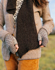 A body shot of Marina wearing a cosy grey cardigan, fingerless mitts and a  dark brown handknit scarf. She is holding the ends of the scarf so that you can see the gentle taper.