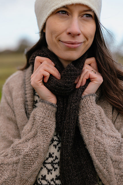 A close up shot of Marina, a white woman with brown hair wearing a cosy grey cardigan, felted hat and a dark brown handknit scarf. She is holding the body of the scarf in her hands and everything looks very warm and cosy