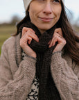 A close up shot of Marina, a white woman with brown hair wearing a cosy grey cardigan, felted hat and a dark brown handknit scarf. She is holding the body of the scarf in her hands and everything looks very warm and cosy