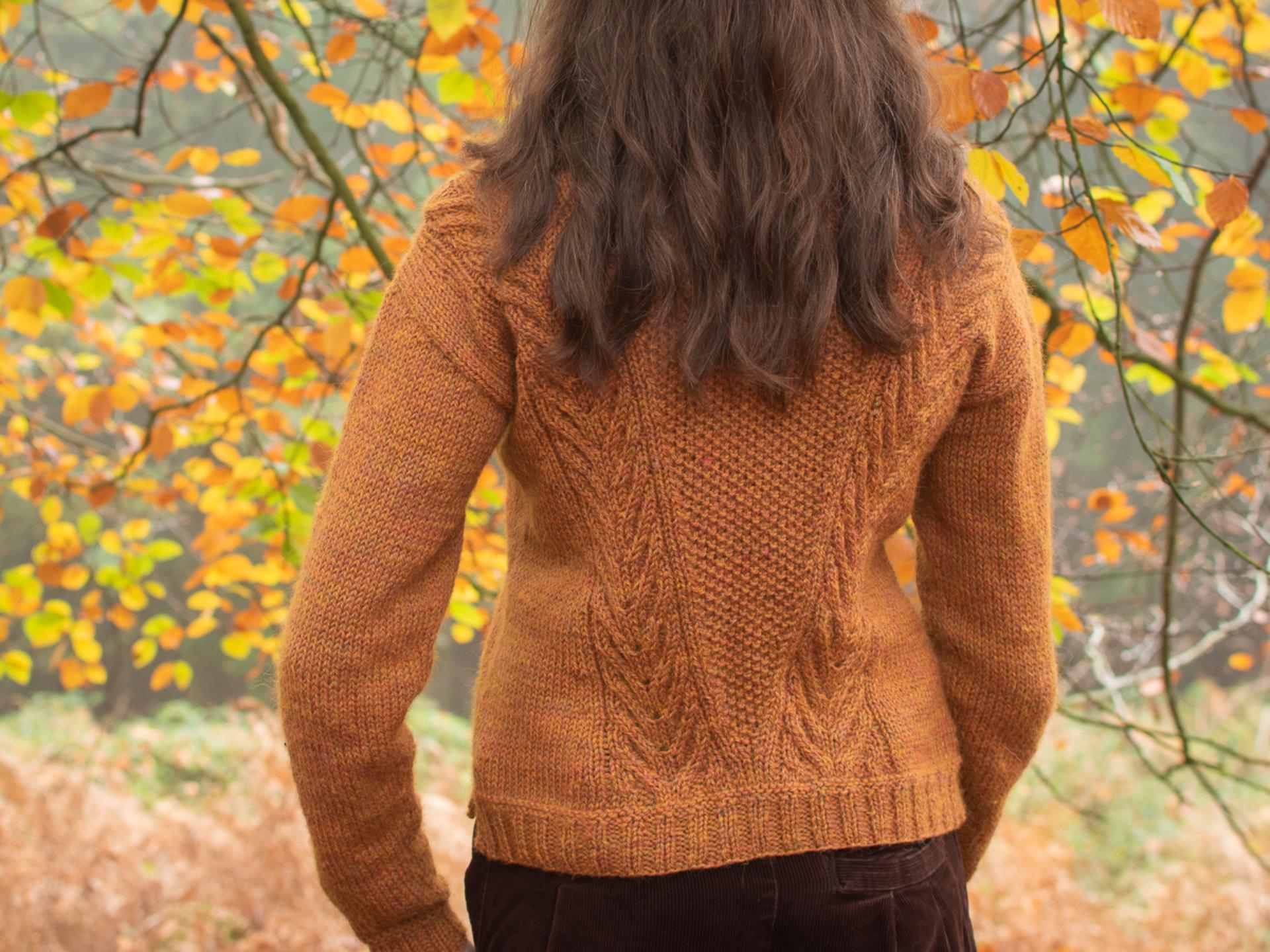 A &#39;from behind&#39; shot of a person outdoors in an autumnal forest setting wearing a rust-coloured, textured handknit sweater.