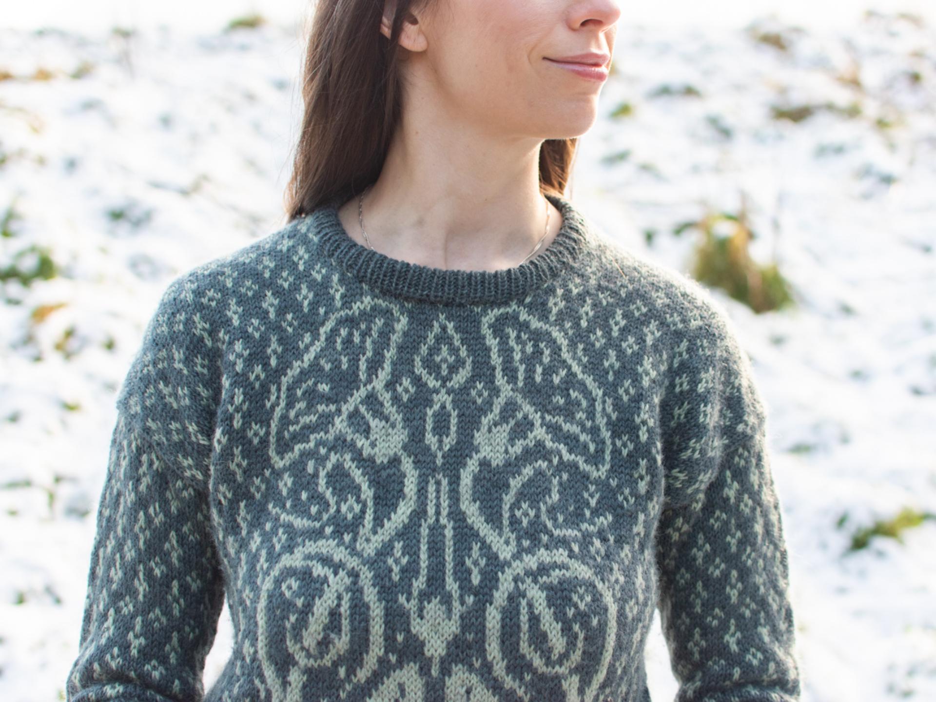 A close-up of Marina, a dark haired white woman wearing a green hand-knitted sweater with detailed botanical colourwork on the chest. The snowy background is softly blurred making the jumper the focus of the image. 