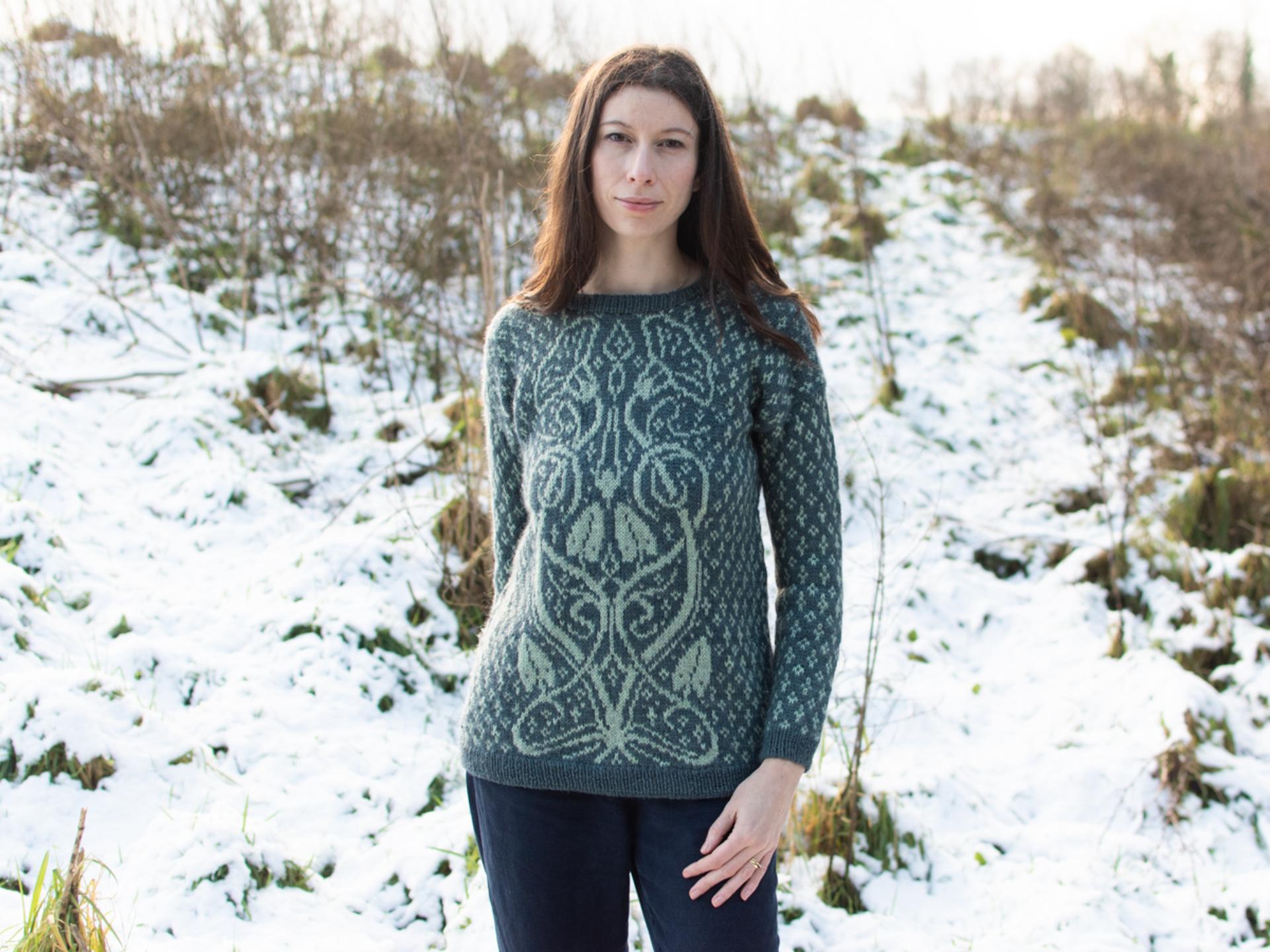 Marina, a dark haired, white woman stands facing the camera in a snowy outdoor setting, wearing a green hand-knitted sweater with an intricate botanical inspired colourwork design on the front. The sweater is paired with dark trousers, and the natural surroundings of snow-covered hills and sparse trees add a wintry charm to the scene.