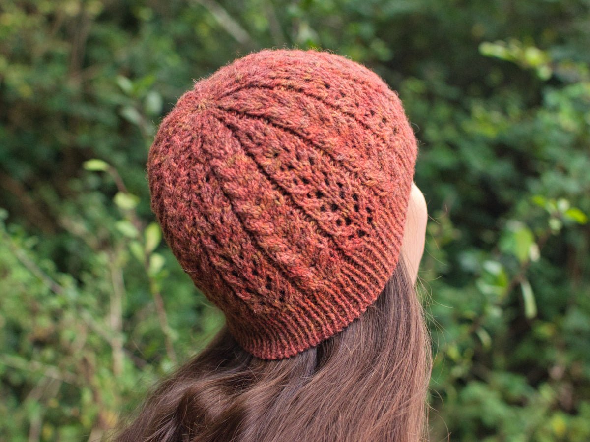 A close-up view of the back of a woman's head. She is wearing a hand knit hat with a lace pattern in warm, autumnal shades of red, orange, and brown. 