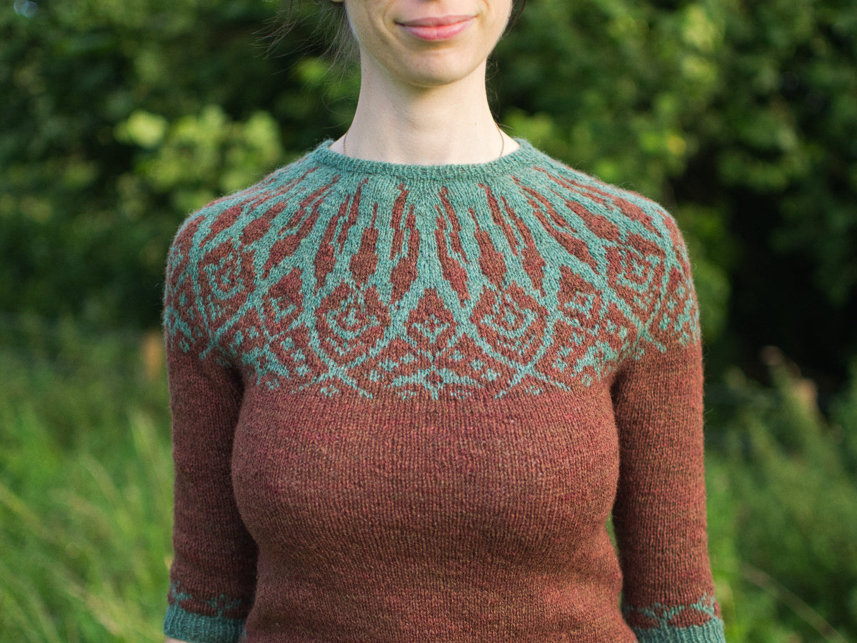 Marina is wearing a knitted sweater made with hand-dyed yarn. It features a colourwork yoke design in earthy shades of brown and teal green. The intricate pattern resembles leaves or floral shapes, creating a rustic, nature-inspired look