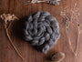 A braided bundle of dark gray British wool fibre resting on a wooden surface, surrounded by dried botanicals such as lavender stems and seed heads