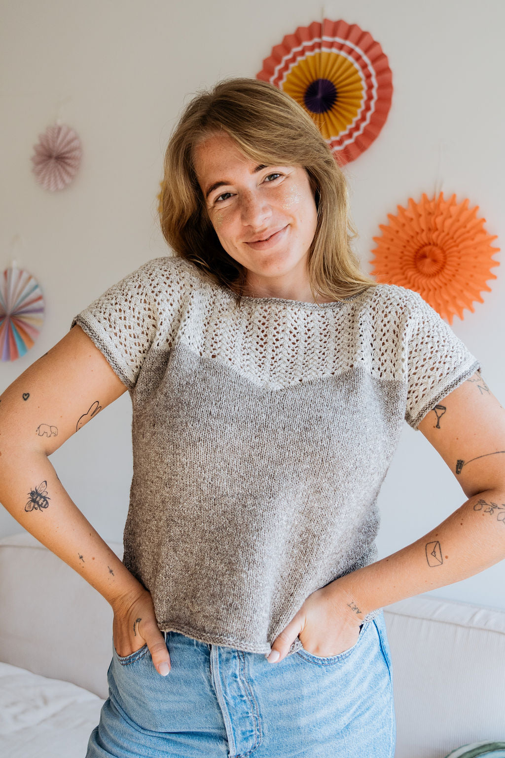 A woman poses in a light, hand knitted top with lace detail, against a backdrop of colorful paper decorations.