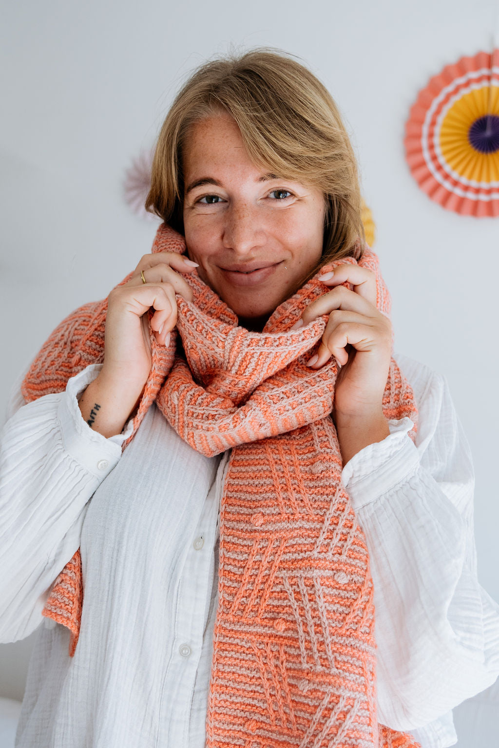 A person wearing a soft, patterned hand knit coral scarf over a white blouse, with decorative paper fans in the background.