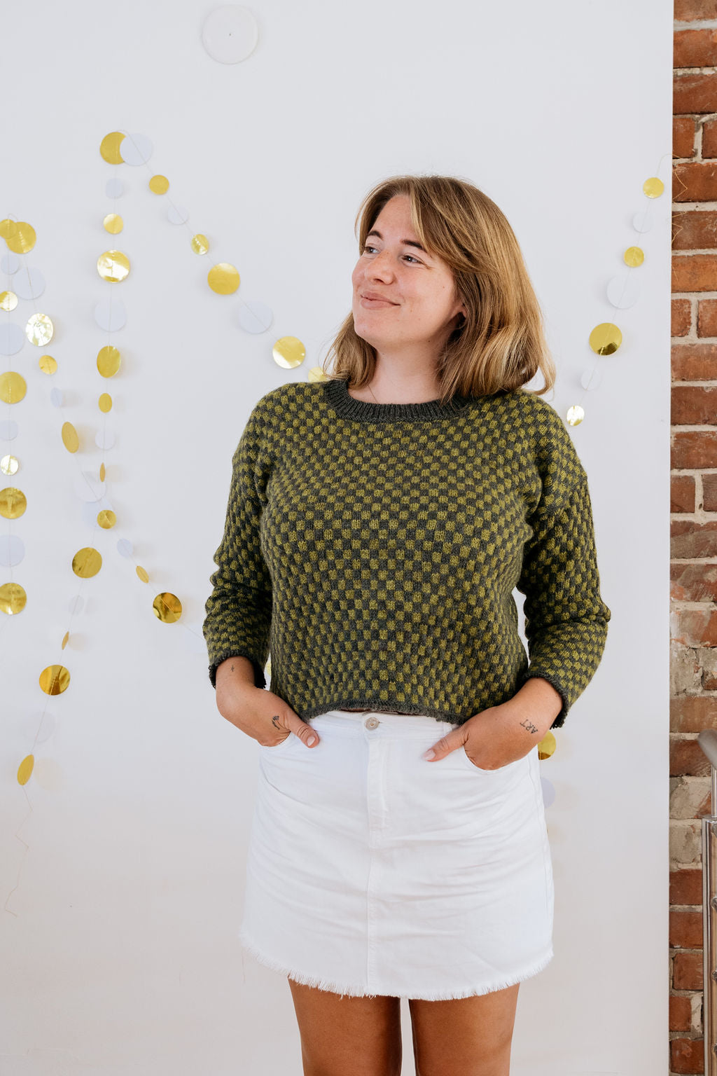 A white person in a green checkered handknit sweater made using hand dyed british wool yarn and white skirt stands smiling in front of a wall with gold decorations.