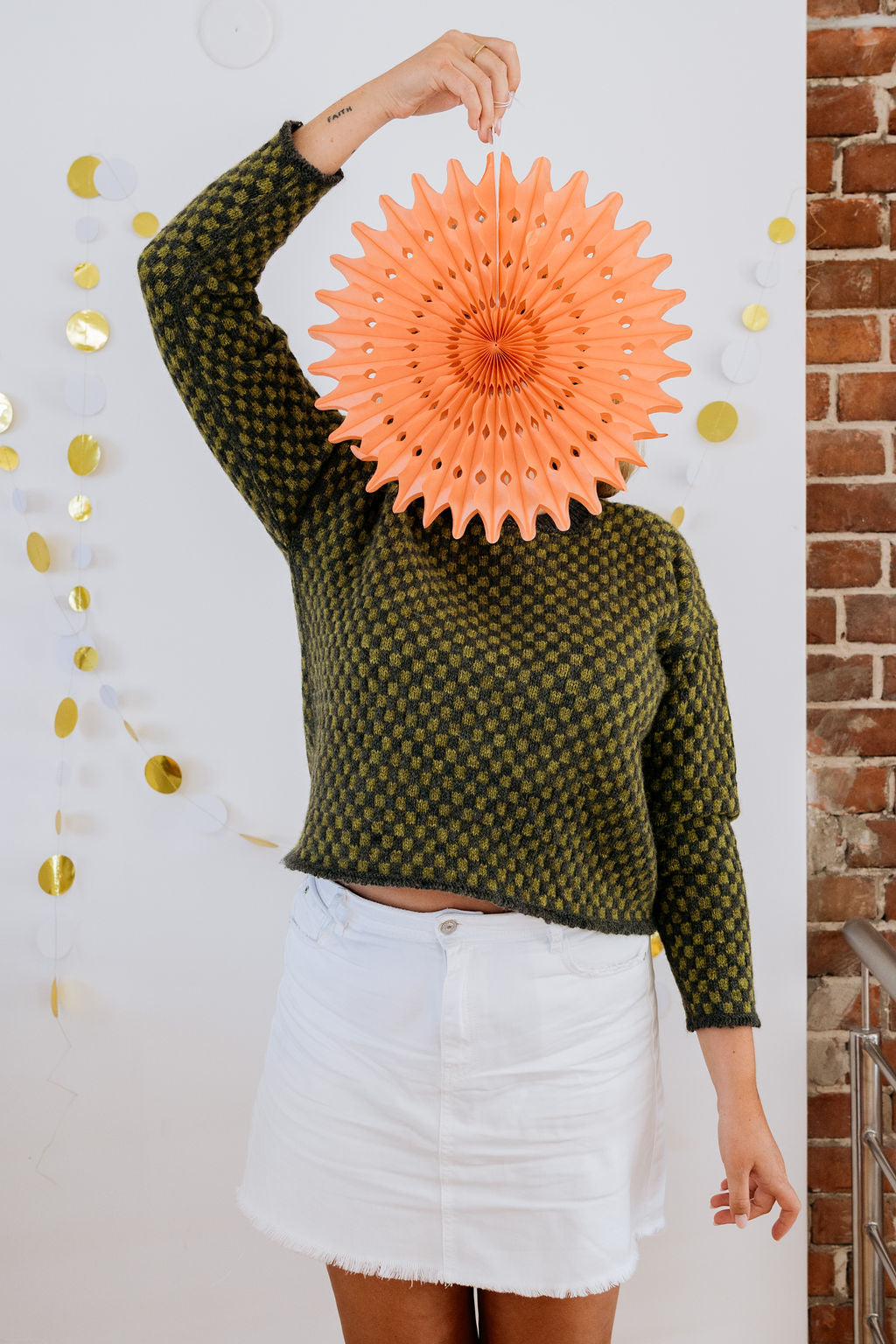 A white person in a green checkered handknit sweater and white skirt is posing for the camera holding a pink paper decoration in front of her face. On the wall behind her are some gold and white paper decorations. 