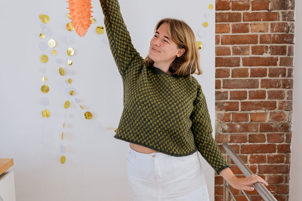 A white person in a green checkered handknit sweater made using hand dyed british wool yarn and white skirt stands smiling in front of a wall with gold decorations. She is holding a paper decoration up in the air with one hand and looking to the side with a smile on her face.