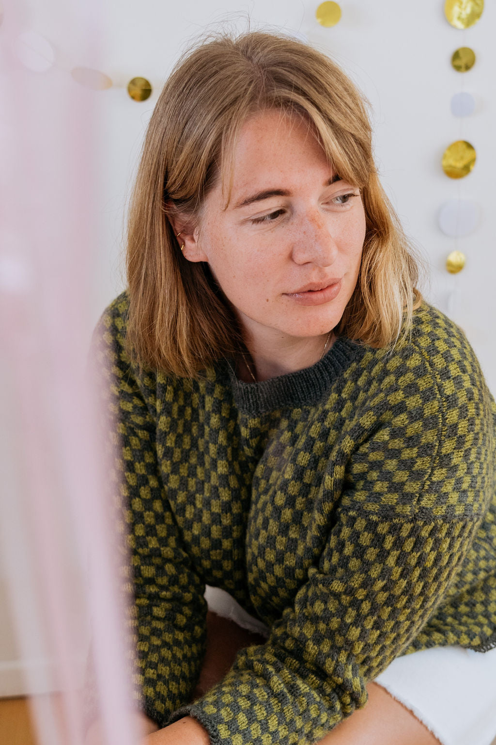 A white person in a green checkered sweater and white skirt sits looking away from the camera. Behind her is a wall with gold decorations.