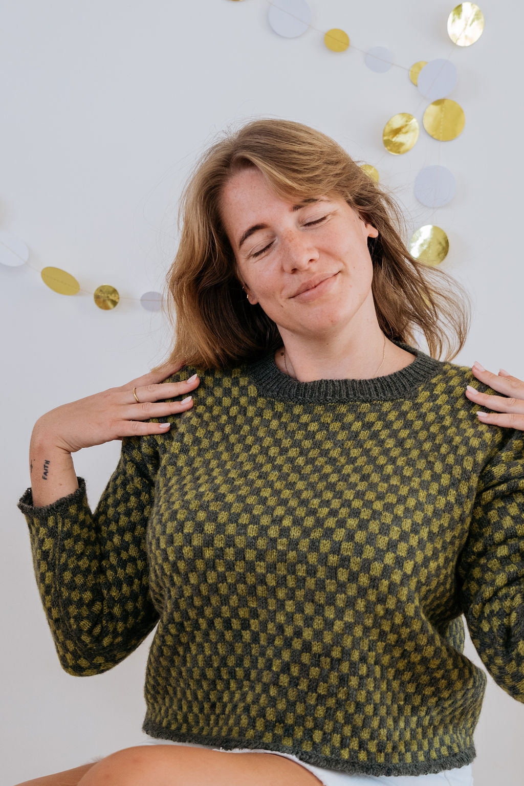 A white person in a green checkered handknit sweater and white skirt is posing and smiling for the camera with her eyes closed and hands on her shoulders. On the wall behind her are some gold and white paper decorations. 