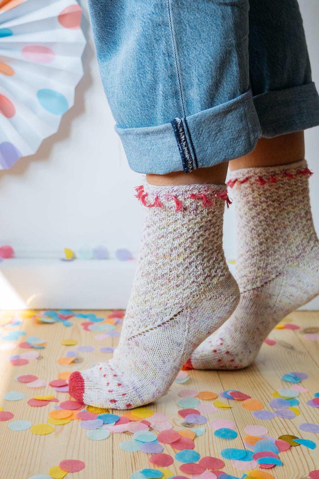 Close up shot of a persons ankles. They are wearing pastel coloured hand knitted socks with pink accents and blue rolled-up jeans, standing on a wooden floor scattered with colourful confetti.