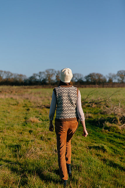 A shot of marina walking through a field. Her back is to the camera and she is wearing a colourwork tank top with branch like details and colours, and a white felted knit hat. 