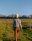 A shot of marina walking through a field. Her back is to the camera and she is wearing a colourwork tank top with branch like details and colours, and a white felted knit hat. 