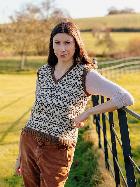 Marina is standing against a countryside fence in a sunny field, wearing a hand-knit, v-neck tank top with intricate stranded colourwork in cream and brown shades. She is looking directly at the camera with a soft smile.