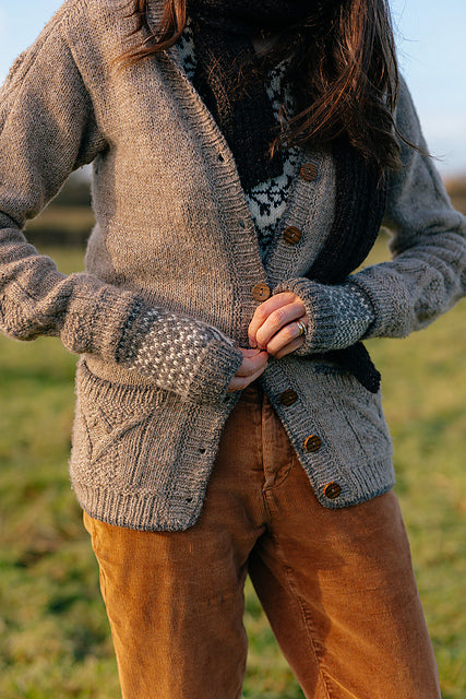A torso shot of Marina who is standing outside wearing a variety of handknit items. The focus is on her buttoning up a grey cardigan wearing grey and white colourwork mitts