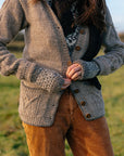 A torso shot of Marina who is standing outside wearing a variety of handknit items. The focus is on her buttoning up a grey cardigan wearing grey and white colourwork mitts