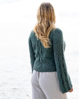 A from behind view of a woman wearing a handknit cardigan. It is made using a teal hand-dyed yarn and features intricate knitted cables along the sleeves and down the back. 