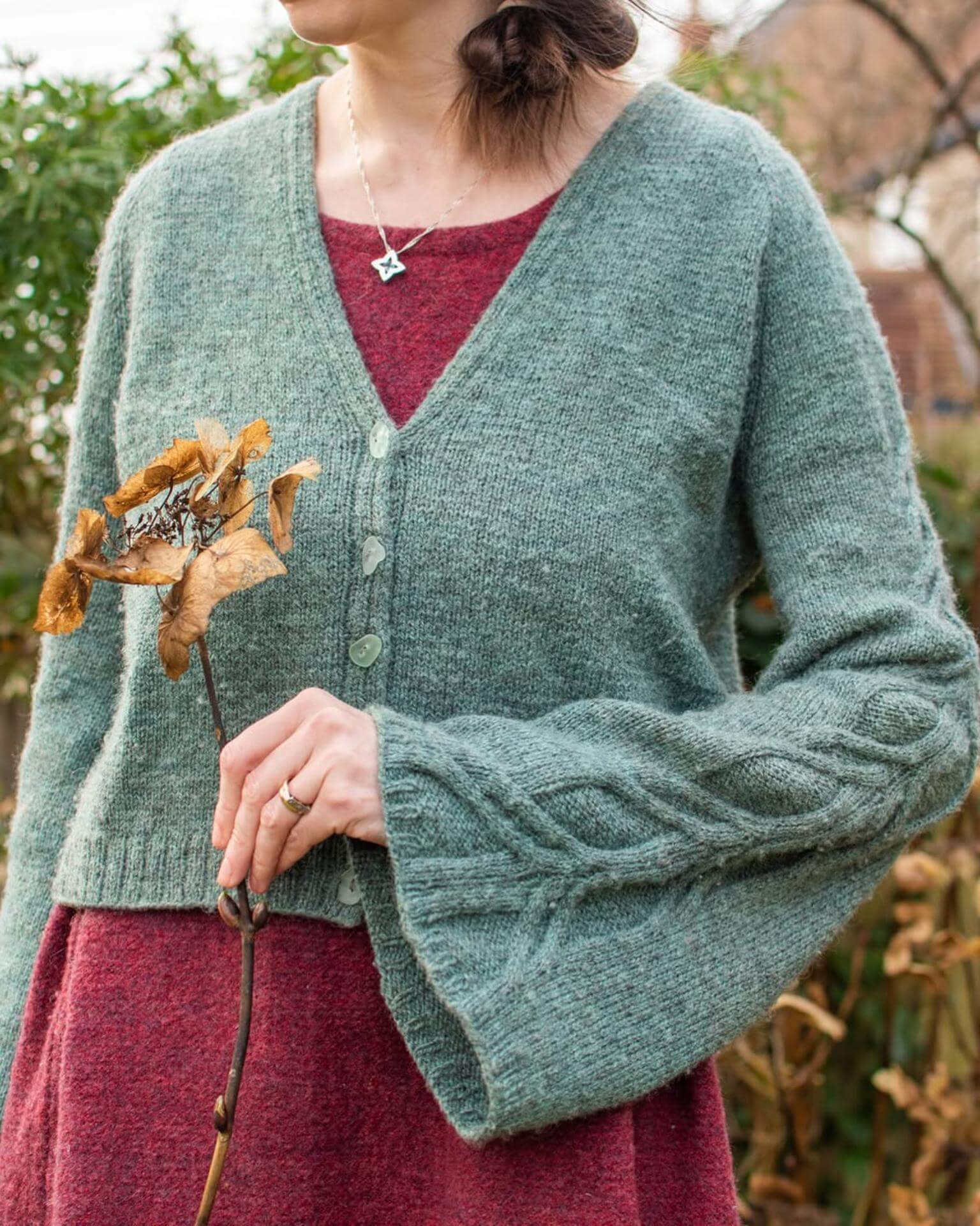 A close up picture of Marina's torso. She is wearing a pink woollen dress with a teal hand knit cardigan over the top, knit using hand dyed British wool yarn. She is holding a stem in her hand and you can see the cabling detail of the wide cardigan sleeve nicely.