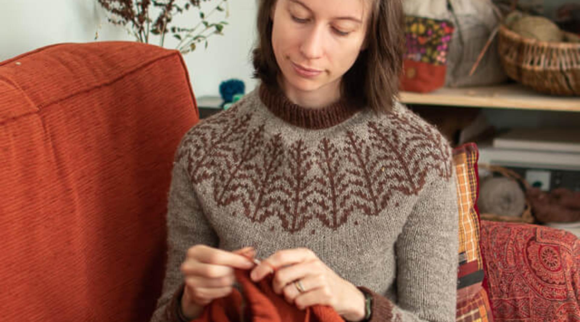 A close up picture of marina sitting knitting with orange yarn on an orange sofa, wearing a hand-knitted jumper